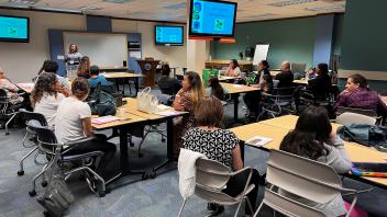 Classroom workshop with attendees at tables listening to a presenter and projection screens.